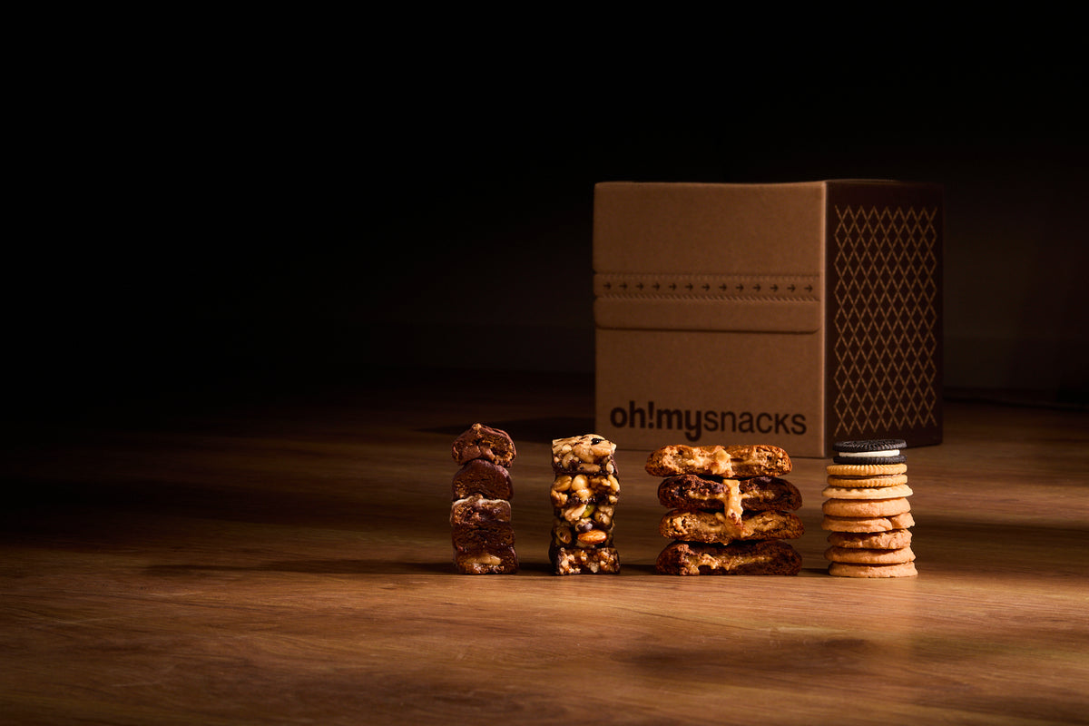 A closed Oh!My box on a wooden table, with stacks of protein balls, nut bars, cakes and biscuits in front of it