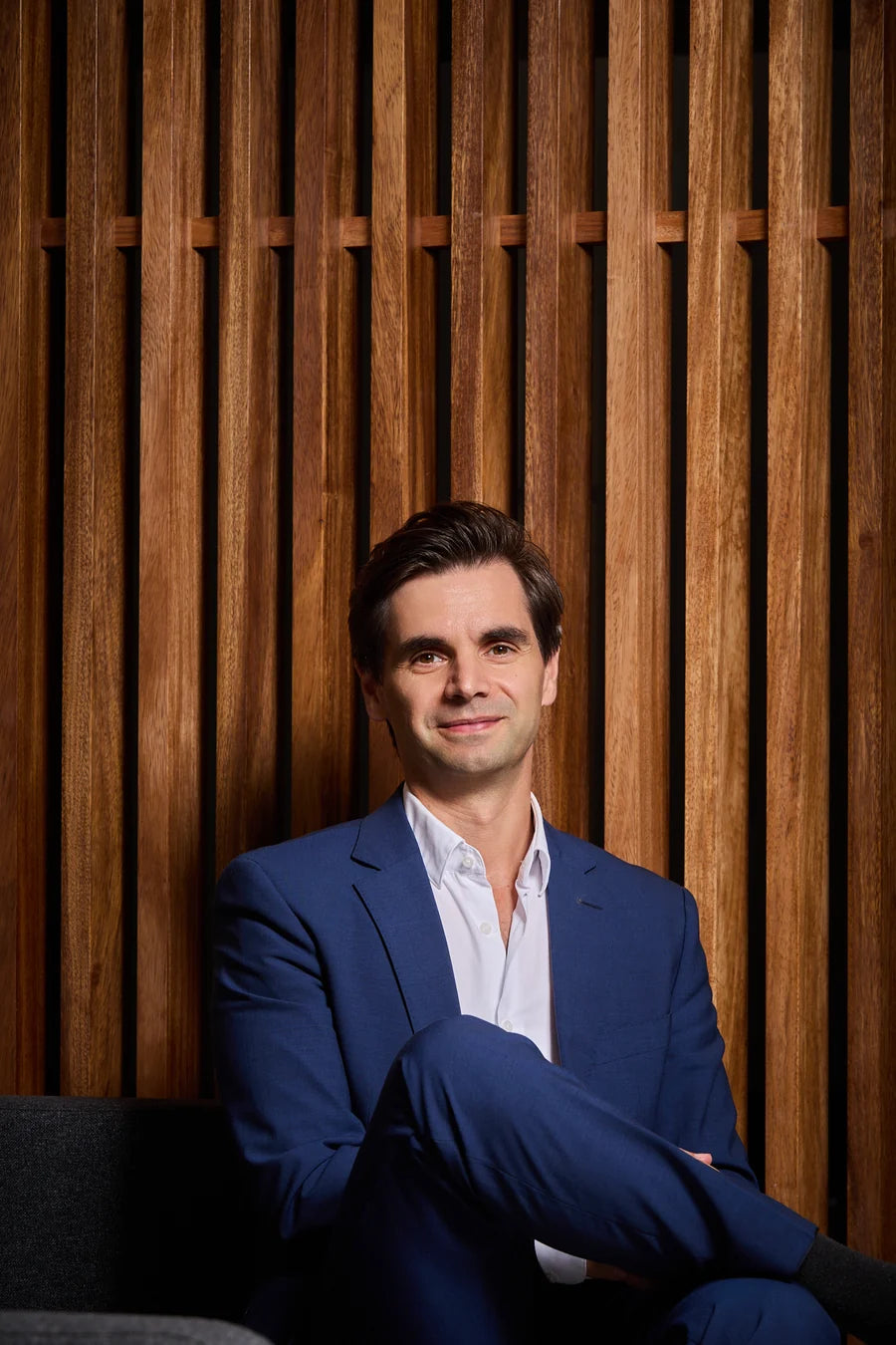 Ricardo Alves, founder of Oh!My Snacks, seated in a blue suit against wooden slats